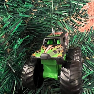 May include: A miniature monster truck ornament with black tires and a green and black body. The truck features a flame design and is suspended by a silver string, perfect for holiday decorating.