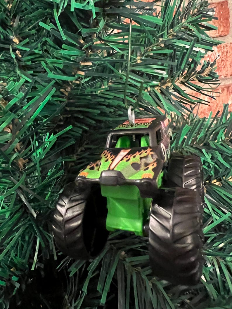 May include: A miniature monster truck ornament with black tires and a green and black body. The truck features a flame design and is suspended by a silver string, perfect for holiday decorating.