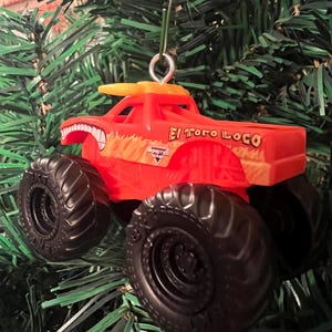 Puede incluir: Adorno navide&ntilde;o de cami&oacute;n monstruo de juguete rojo y naranja. El cami&oacute;n tiene neum&aacute;ticos negros grandes y las palabras "El Toro Loco" en el lateral. El adorno cuelga de un &aacute;rbol de Navidad verde.