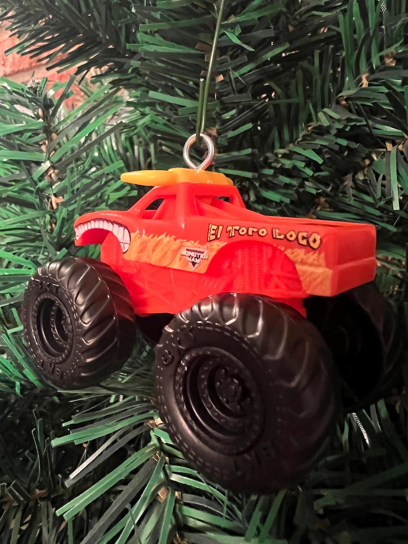 Puede incluir: Adorno navide&ntilde;o de cami&oacute;n monstruo de juguete rojo y naranja. El cami&oacute;n tiene neum&aacute;ticos negros grandes y las palabras "El Toro Loco" en el lateral. El adorno cuelga de un &aacute;rbol de Navidad verde.