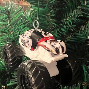 May include: A white and black monster truck ornament with large black tires. The truck features a Dalmatian dog design on the cab and is suspended by a silver wire loop, perfect for holiday decorating.