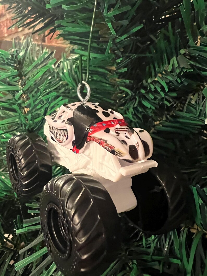 May include: A white and black monster truck ornament with large black tires. The truck features a Dalmatian dog design on the cab and is suspended by a silver wire loop, perfect for holiday decorating.