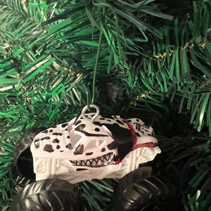 May include: A white and black monster truck Christmas ornament with black tires. The truck has a Dalmatian dog design with a red collar and a menacing grin. The ornament hangs from a silver metal loop.