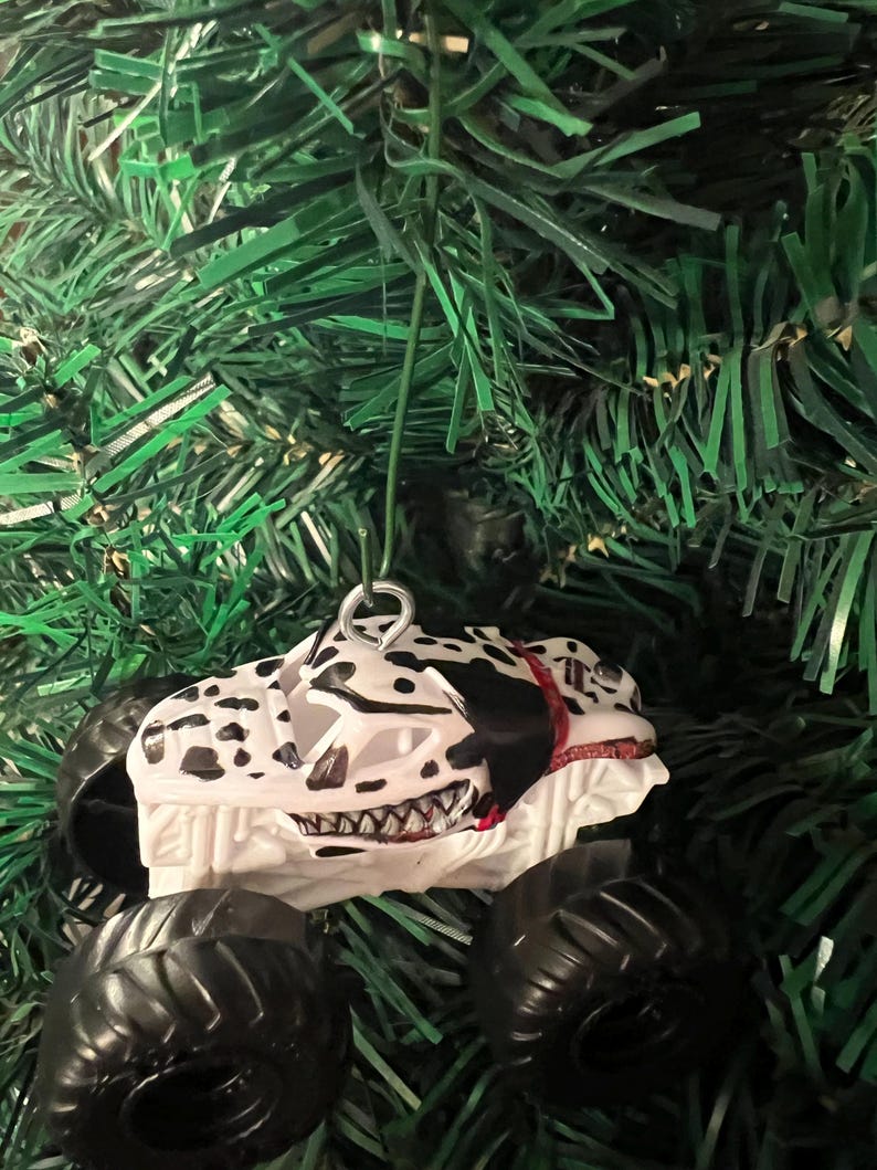 May include: A white and black monster truck Christmas ornament with black tires. The truck has a Dalmatian dog design with a red collar and a menacing grin. The ornament hangs from a silver metal loop.