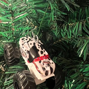 May include: A white and black monster truck Christmas ornament with a Dalmatian dog design. The ornament has large black tires and a red collar. The ornament is hanging on a green Christmas tree.