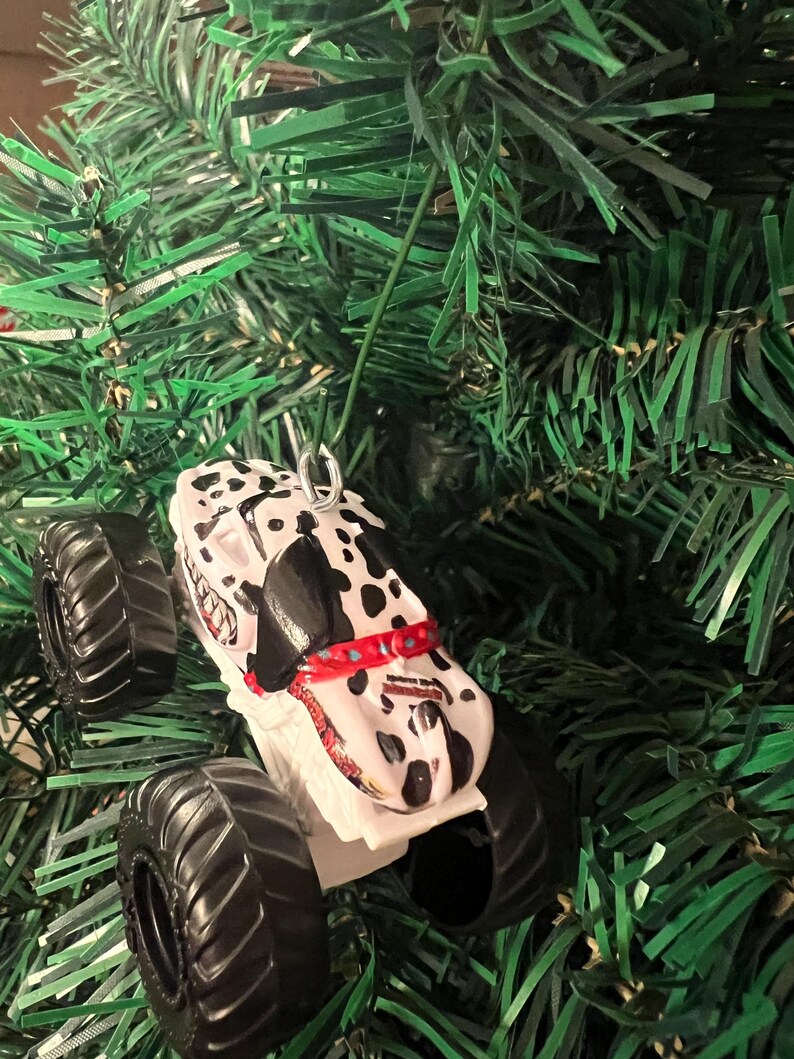 May include: A white and black monster truck Christmas ornament with a Dalmatian dog design. The ornament has large black tires and a red collar. The ornament is hanging on a green Christmas tree.