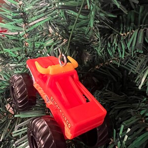 Puede incluir: Adorno navide&ntilde;o de cami&oacute;n monstruo de juguete rojo con neum&aacute;ticos negros y cuernos de toro amarillos. El texto "El Toro Loco" est&aacute; impreso en el lateral. Colgando de un &aacute;rbol de Navidad verde.