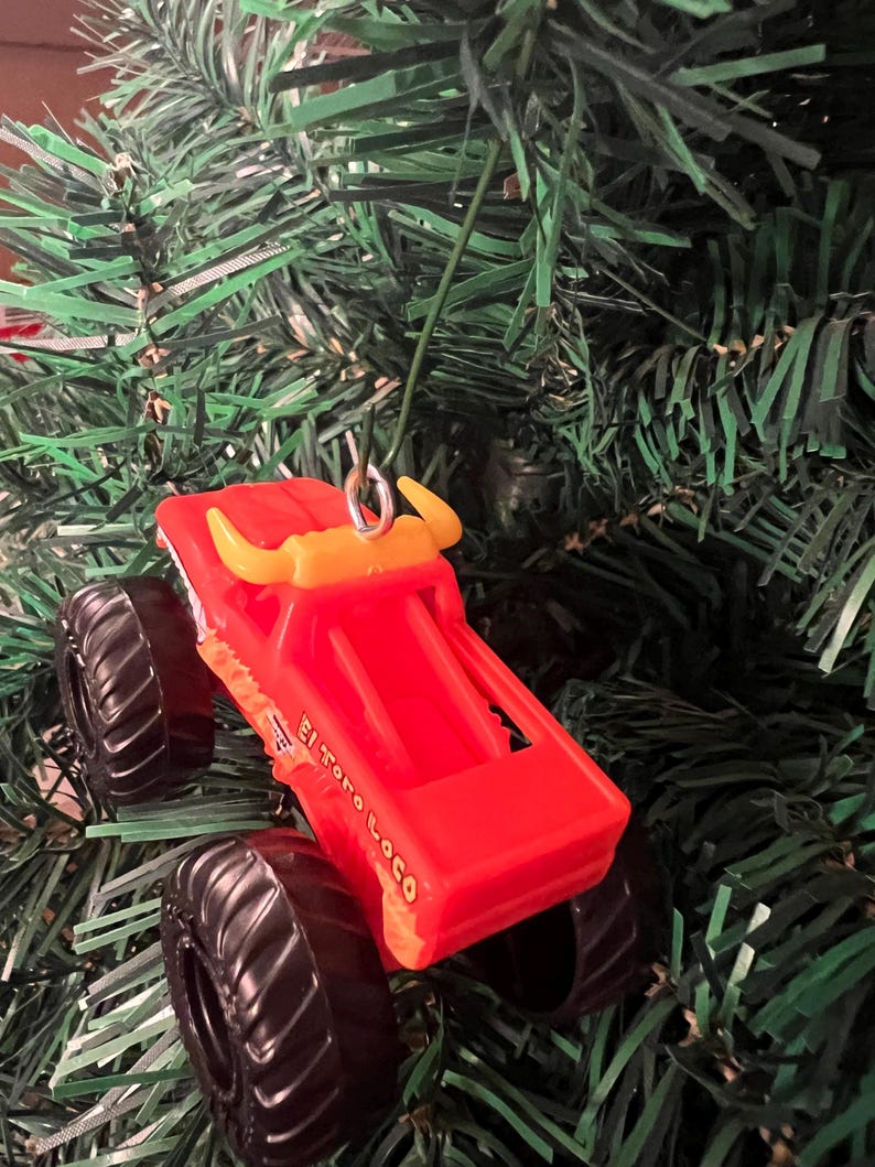 Puede incluir: Adorno navide&ntilde;o de cami&oacute;n monstruo de juguete rojo con neum&aacute;ticos negros y cuernos de toro amarillos. El texto "El Toro Loco" est&aacute; impreso en el lateral. Colgando de un &aacute;rbol de Navidad verde.