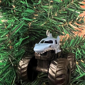 May include: A shark-themed monster truck Christmas ornament. The truck is light blue with a shark fin and mouth design, black tires, and a silver hanger. The ornament is displayed on a green Christmas tree.