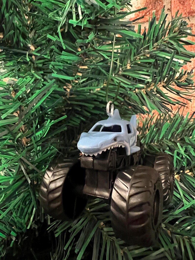 May include: A shark-themed monster truck Christmas ornament. The truck is light blue with a shark fin and mouth design, black tires, and a silver hanger. The ornament is displayed on a green Christmas tree.