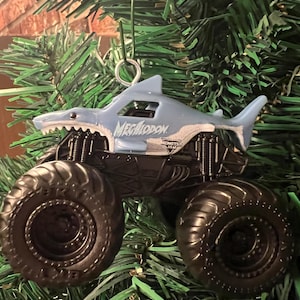 May include: A light blue Megalodon monster truck ornament with black wheels and a shark-shaped body. The ornament is suspended by a silver metal loop and is set against a backdrop of green pine needles.