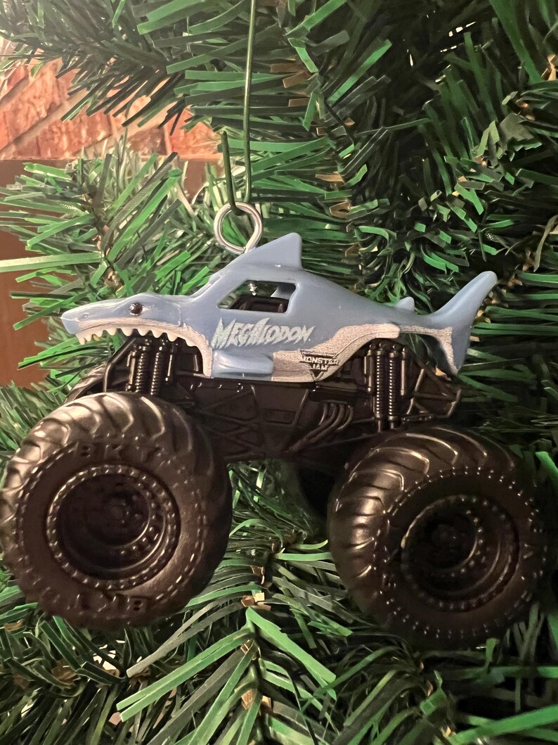 May include: A light blue Megalodon monster truck ornament with black wheels and a shark-shaped body. The ornament is suspended by a silver metal loop and is set against a backdrop of green pine needles.