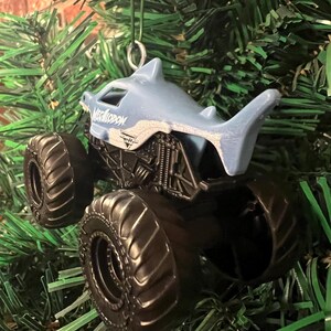 May include: A Megalodon monster truck ornament with a shark-shaped cab and large black tires. The ornament is suspended against a backdrop of green Christmas tree branches. The text "Megalodon" is visible on the cab.