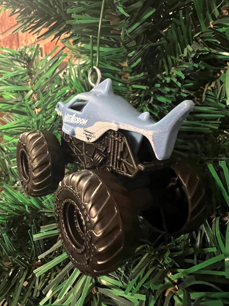 May include: A Megalodon monster truck ornament with a shark-shaped cab and large black tires. The ornament is suspended against a backdrop of green Christmas tree branches. The text "Megalodon" is visible on the cab.