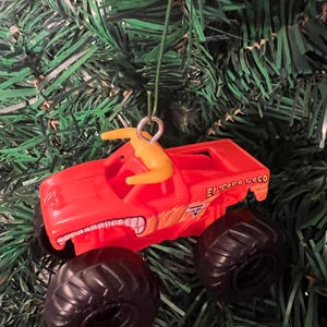 Puede incluir: Adorno navide&ntilde;o de cami&oacute;n monstruo de juguete rojo con neum&aacute;ticos negros y la inscripci&oacute;n "El Toro Loco" en el lateral. El adorno tiene una figura amarilla en la parte superior y cuelga de una cuerda verde.
