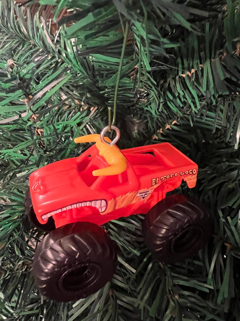 Puede incluir: Adorno navide&ntilde;o de cami&oacute;n monstruo de juguete rojo con neum&aacute;ticos negros y la inscripci&oacute;n "El Toro Loco" en el lateral. El adorno tiene una figura amarilla en la parte superior y cuelga de una cuerda verde.