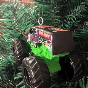 May include: A Grave Digger monster truck ornament with a black top and green chassis. The truck has large black tires and the words "Grave Digger" are visible on the side. The ornament is hanging from a green Christmas tree.