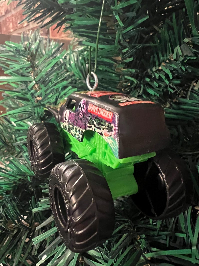 May include: A Grave Digger monster truck ornament with a black top and green chassis. The truck has large black tires and the words "Grave Digger" are visible on the side. The ornament is hanging from a green Christmas tree.