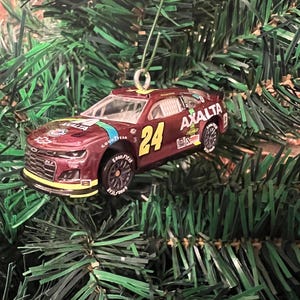 May include: A maroon Chevrolet Camaro ZL1 race car ornament featuring the number 24 in yellow, the Axalta logo in white, and Goodyear branding on the black tires. The vehicle has a silver hanging loop attached to the roof for display purposes.