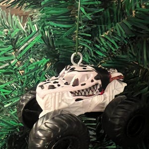 May include: A white and black monster truck ornament with a dinosaur skull design. The ornament has large black tires and a red interior. It is hanging from a green Christmas tree.