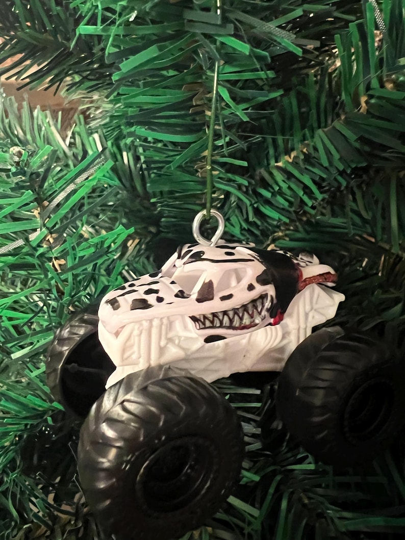 May include: A white and black monster truck ornament with a dinosaur skull design. The ornament has large black tires and a red interior. It is hanging from a green Christmas tree.