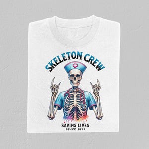 May include: White t-shirt with a blue and white skeleton wearing a nurse's hat and holding up two fingers in a rock and roll hand gesture. The text "Skeleton Crew" is in a blue circle above the skeleton and the text "Saving Lives Since 1853" is below the skeleton.