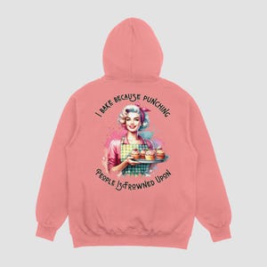 Pu&ograve; includere: Felpa con cappuccio rosa con una stampa grafica di una donna che tiene un vassoio di cupcake. Il testo sulla felpa recita "I bake because punching people is frowned upon."