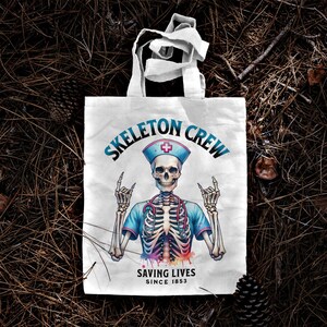 May include: White tote bag with a graphic of a skeleton wearing a nurse's uniform and a stethoscope. The text "Skeleton Crew Saving Lives Since 1853" is printed on the bag.