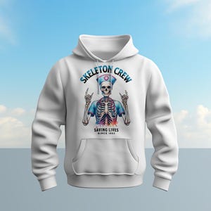 May include: White hoodie with a graphic of a skeleton wearing a nurse's uniform and holding a stethoscope. The text "Skeleton Crew Saving Lives Since 1853" is printed on the hoodie.