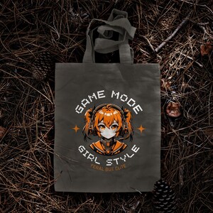 May include: A gray canvas tote bag with a graphic design featuring an anime girl wearing headphones and the text "GAME MODE GIRL STYLE FERAL BUT CUTE".
