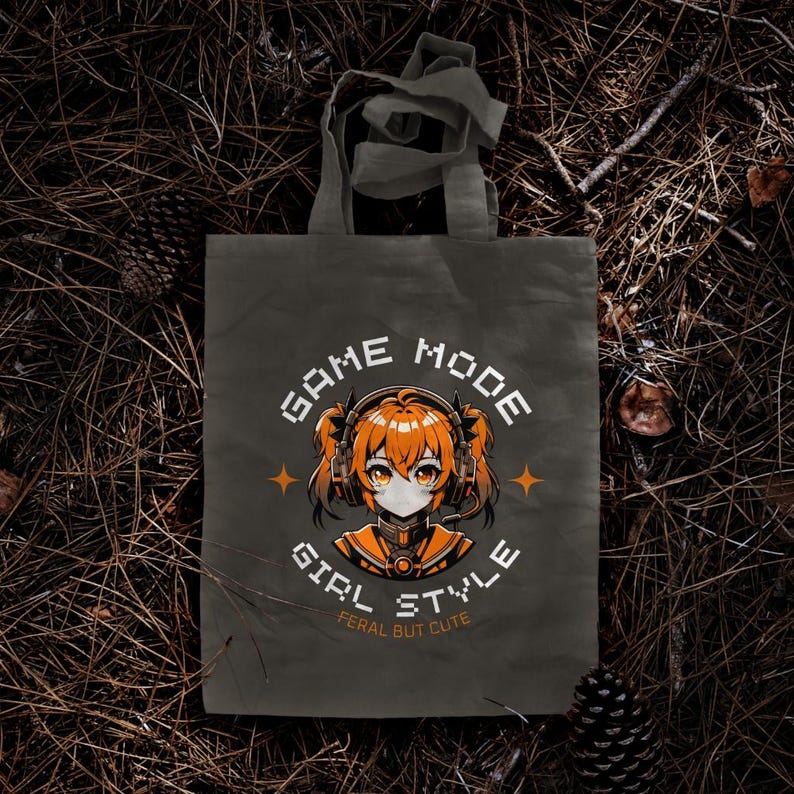 May include: A gray canvas tote bag with a graphic design featuring an anime girl wearing headphones and the text "GAME MODE GIRL STYLE FERAL BUT CUTE".
