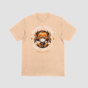 May include: A light beige t-shirt with a graphic print of a girl with orange hair wearing headphones and a black mask. The text "GAME MODE GIRL STYLE" is printed in black and orange on the shirt. The text "FERAL BUT CUTE" is printed in black below the graphic.