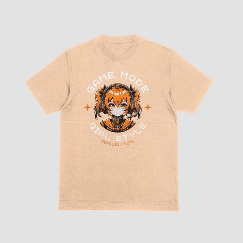 May include: A light beige t-shirt with a graphic print of a girl with orange hair wearing headphones and a black mask. The text "GAME MODE GIRL STYLE" is printed in black and orange on the shirt. The text "FERAL BUT CUTE" is printed in black below the graphic.