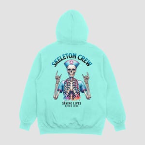 May include: A turquoise blue hooded sweatshirt with a skeleton wearing a nurse's uniform and a red cross. The text "Skeleton Crew Saving Lives Since 1853" is printed on the front.