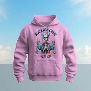 May include: Pink hoodie with a skeleton wearing a nurse's uniform and a stethoscope. The text on the hoodie says "Skeleton Crew Saving Lives Since 1853".