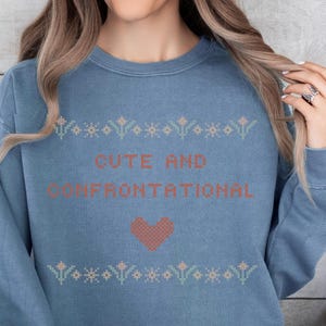 May include: A blue sweatshirt with the text "CUTE AND CONFRONTATIONAL" in orange, along with a heart graphic. The sweatshirt has a crew neck and long sleeves. The design includes floral and snowflake patterns.