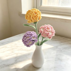 Könnte beinhalten: Drei gehäkelte Blumen in Gelb, Helllila und Rosa, jede mit einem grünen Stiel und Blättern, arrangiert in einer kleinen weißen Keramikvase. Die Blumen weisen komplizierte, gerüschte Blütenblattstrukturen aus Garn auf und stehen aufrecht.
