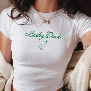 May include: White t-shirt with the text "Lucky Duck" in green script. A green duck graphic holding a shamrock is below the text. The shirt has short sleeves and a crew neck.