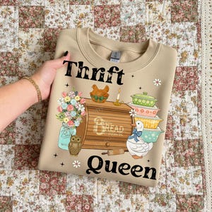 Op de afbeelding: Beige crewneck sweatshirt met een kleurrijke afbeelding. De afbeelding toont de woorden "Thrift Bread Queen" met illustraties van een kip, bloemen, een uil, een eend en gestapelde kommen. De sweatshirt wordt weergegeven op een quilt met bloemenpatroon.