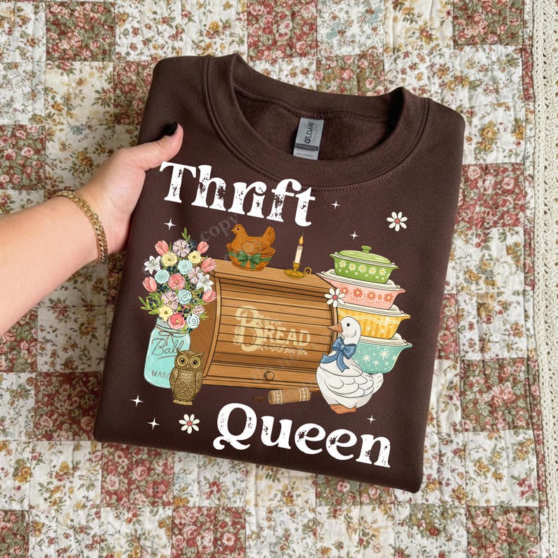 Op de afbeelding: Een bruine sweatshirt met de witte tekst "Thrift Queen". De afbeelding bevat een broodtrommel, een stapel kommen, een pot bloemen, een kip, een uil en een eend.
