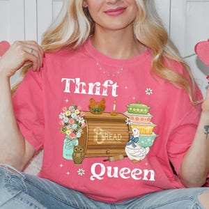 Op de afbeelding: Een roze t-shirt met de witte tekst "Thrift Queen". Het shirt heeft een afbeelding van een broodtrommel, opgestapelde kommen, een pot met bloemen en een uil. Extra afbeeldingen zijn een eend, een kaars en een kip.