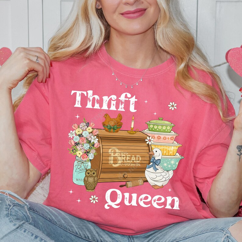 Op de afbeelding: Een roze t-shirt met de witte tekst "Thrift Queen". Het shirt heeft een afbeelding van een broodtrommel, opgestapelde kommen, een pot met bloemen en een uil. Extra afbeeldingen zijn een eend, een kaars en een kip.