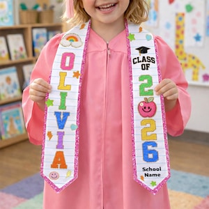 May include: A pink graduation gown with a personalized sash. The sash is white with the name "OLIVIA" in colorful letters and "CLASS OF 2026" with an apple graphic. The sash has a glittery pink border and decorative stickers.