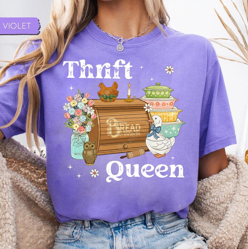 Op de afbeelding: Een paars T-shirt met de tekst "Thrift Queen" en een grafisch ontwerp. Het ontwerp bevat een broodtrommel, bloemen in een pot, een uil, een eend en opgestapelde schalen.