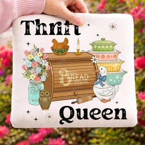 Op de afbeelding: Een wit kussen met de woorden "Thrift Queen" en diverse illustraties. De illustraties omvatten een broodtrommel, een eend met een blauwe strik, een pot bloemen en een stapel kleurrijke ovenschalen.