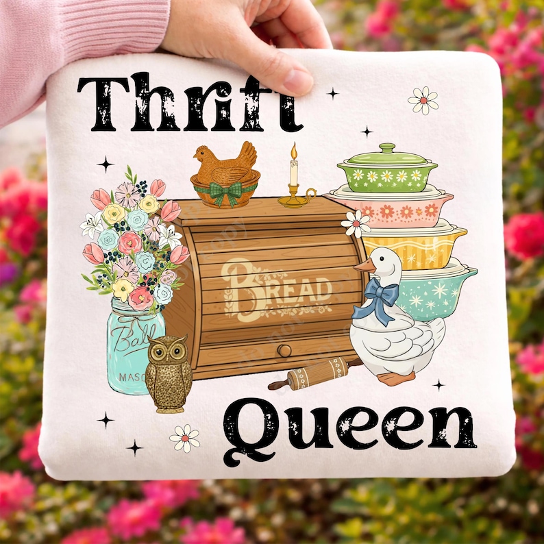 Op de afbeelding: Een wit kussen met de woorden "Thrift Queen" en diverse illustraties. De illustraties omvatten een broodtrommel, een eend met een blauwe strik, een pot bloemen en een stapel kleurrijke ovenschalen.