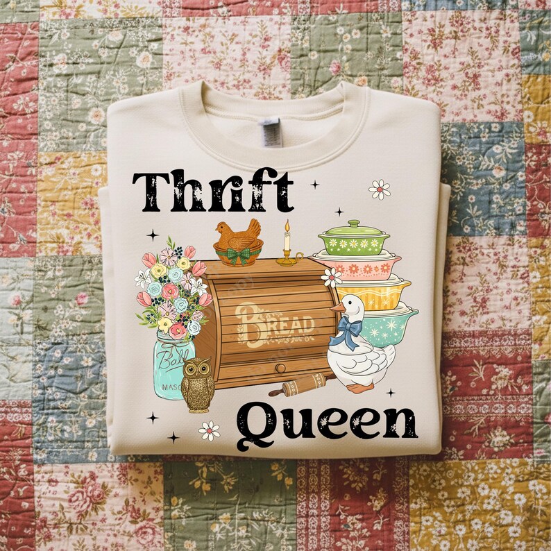 Op de afbeelding: Cr&egrave;mekleurige sweatshirt met de woorden "Thrift Queen" in het zwart. De afbeelding bevat een broodtrommel, gestapelde kommen, een eend, bloemen en een uil. Het ontwerp heeft een vintage, boerderij-esthetiek.