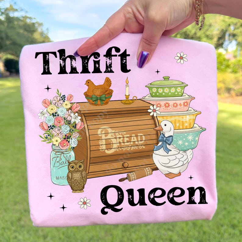 Op de afbeelding: Roze t-shirt met de tekst "Thrift Queen" en illustraties van een broodtrommel, ovenschalen, een eend en bloemen. De afbeelding bevat ook een weckpot, een uil en een deegroller.