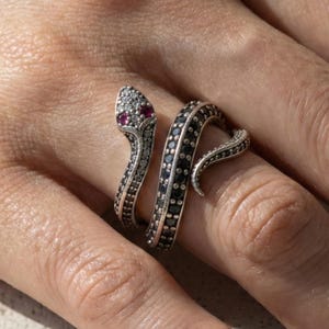 Könnte beinhalten: Ein silberner Schlangenring mit schwarzen und klaren Steinen. Der Schlangenkopf hat zwei rote Steine als Augen. Der Ring umschlingt den Finger und schafft ein einzigartiges und stilvolles Accessoire.