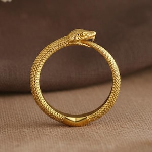 May include: A gold ring depicting an ouroboros, a snake consuming its own tail. The ring has a textured finish and is set against a neutral backdrop, with dried flowers in the background.
